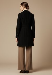 Vrouw met donker haar in een knot, gekleed in een zwarte lange jas, beige wijde broekspijpen en zwarte schoenen met hakken, staande tegen een beige achtergrond.