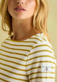 Weißes Oberteil mit horizontalen goldenen Streifen, mit einem runden Halsausschnitt und einem Seitenschild. Glatte Textur, lässiges Design.