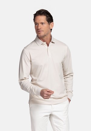 Man in beige lange mouwen poloshirt en witte broek, staand met één hand in de zak en de andere iets omhoog, kijkend naar rechts.