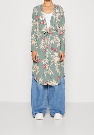 Personne portant un long cardigan vert à motif floral noué à la taille, un jean large bleu, des chaussures blanches et un haut blanc en dessous.