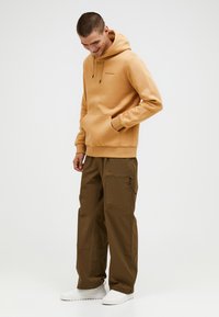 Beige hoodie met een kangoeroezak en een drawstring, gecombineerd met olijfgroene cargobroeken met ritszakken, afgewerkt met witte schoenen.