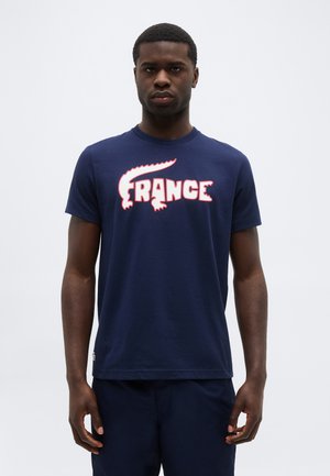 Νεαρός άνδρας που στέκεται και κοιτάει μπροστά, φορώντας μπλε ναυτικό μπλουζάκι με ένα στυλιζαρισμένο λογότυπο "FRANCE" σε λευκό και κόκκινο χρώμα στο στήθος.