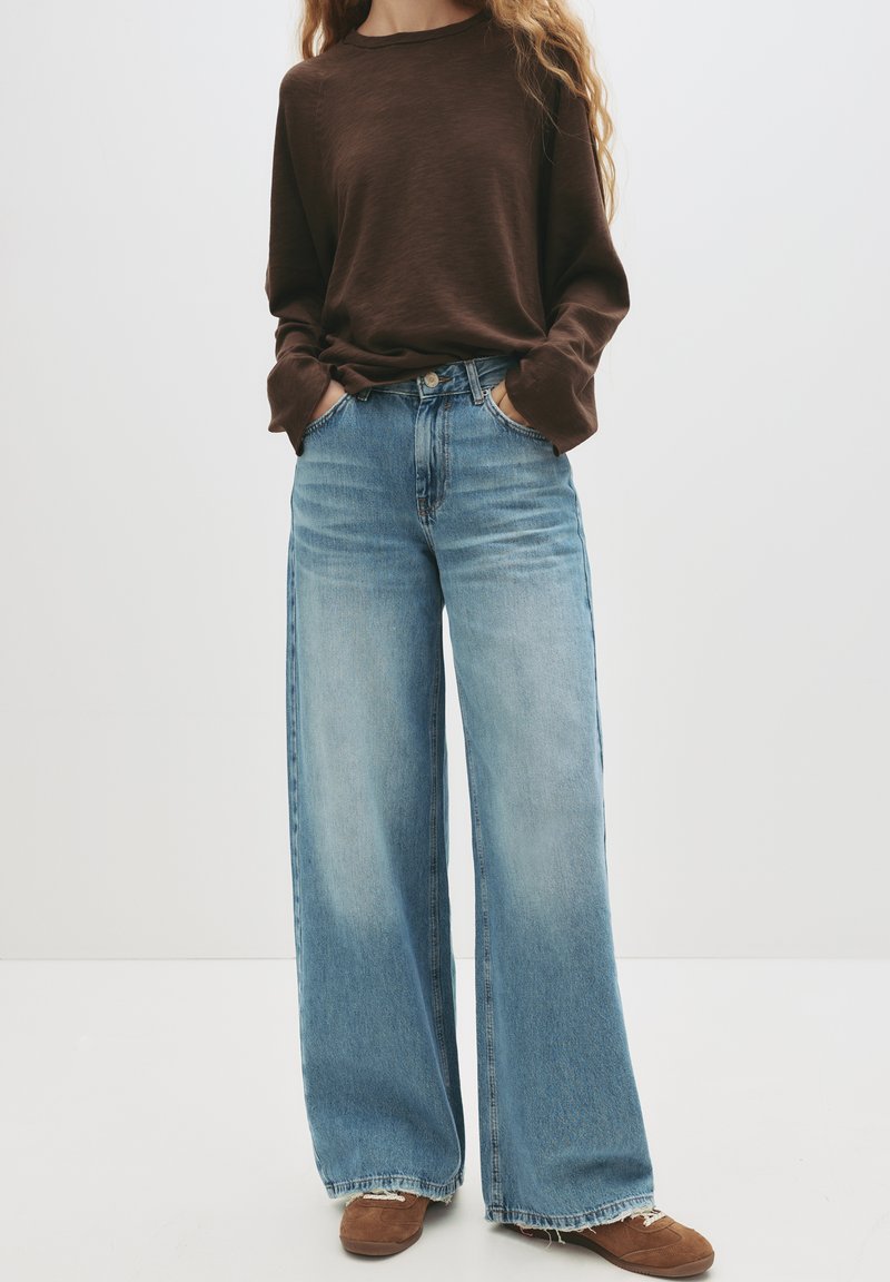 Rjav dolg rokav top, kombiniran s svetlo modrimi širokimi kavbojkami. Kavbojke imajo zbledelo pranje in strgan rob, dopolnjene z rjavimi čevlji.