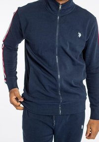 Marinblå zip-up träningsoveralljacka tillverkad av mjukt tyg, med röda och vita ränder längs ärmarna och en liten logotyp på bröstet.