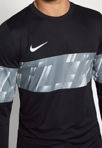 Czarna sportowa koszulka z długim rękawem z szarym paskiem graficznym na klatce piersiowej z kanciastymi wzorami oraz białym logo Nike.