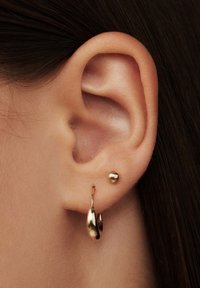 Goldene Creolen in Tropfenform mit glatter Oberfläche, kombiniert mit einem kleinen goldenen Ballstecker, vor einem hellen Hautton.