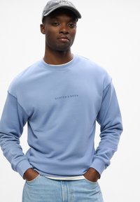 Sudadera azul claro con puños acanalados, que lleva el logo "SCOTCH & SODA", usada sobre una camisa blanca, combinada con jeans de denim claro.
