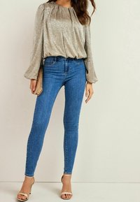 Blouse longue à manches pleats brillants en argent, associée à un jean bleu ajusté orné de strass éparpillés et des talons dorés ouverts.