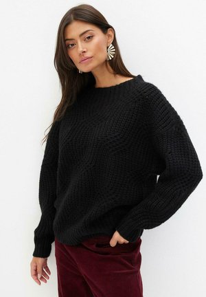 Schwarzer Strickpullover mit strukturiertem Muster, Rundhalsausschnitt und langen Ärmeln, kombiniert mit kastanienbraunen Cordhosen.