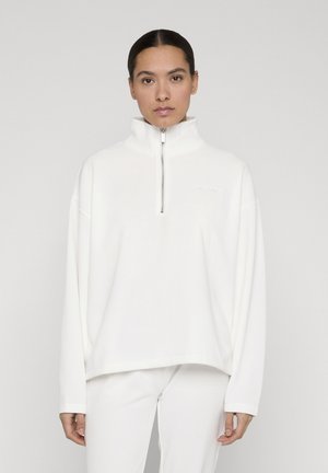 Tummatukkainen nainen, jolla on matala nuttura, pukeutunut valkoiseen, korkeakauluksiseen vetoketjuneuleeseen ja valkoisiin housuihin yksinkertaista harmaata taustaa vasten.