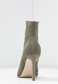 Bottine à talon haut vert olive avec un dessus en maille texturée et un talon aiguille lisse de couleur claire. Fermeture éclair à l'arrière.