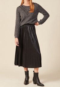 Grauer Pullover mit Perlenakzenten und langen Ärmeln, kombiniert mit einem schwarzen Faltenrock und schwarzen Stiefeletten, vor einem neutralen Hintergrund.