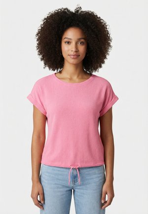 Femme aux cheveux bouclés portant un haut rose à manches courtes avec les poignets retroussés et un jean bleu clair, debout devant un fond blanc.