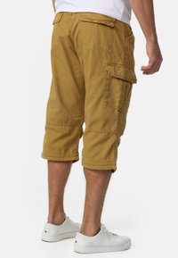 Bermudas cargo jusqu'aux genoux en coton beige, avec plusieurs poches latérales, des détails cousus et une coupe décontractée. Portées avec des baskets blanches.