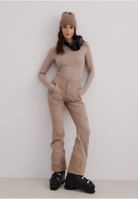 Beige langermet topp kombinert med lysebeige skibukser med sidelommer, elastisk midje og svarte skistøvler for vintersport.