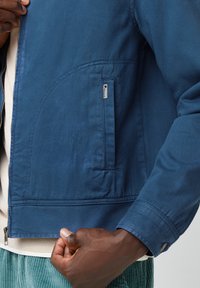 Blauwe jas van gladde stof, met een voorschuif, een zijzak en geribbelde manchetten. Opvallend logo tag op de zak.