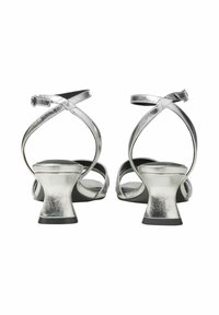 Silvriga remsandaler med hög klack har en metallisk yta, spänne vid vristen och en flaread klack med en svart sula.