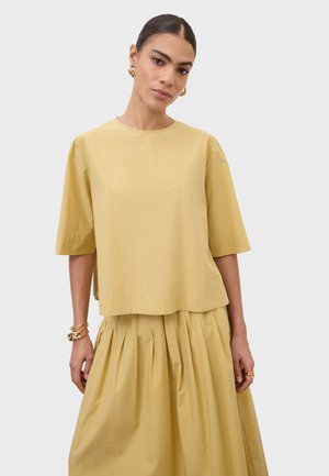 Kobieta z ciemnymi włosami związanymi do tyłu, ubrana w luźny beżowy top z krótkim rękawem i plisowaną beżową spódnicę, uzupełniona złotymi kolczykami kołami i bransoletkami.