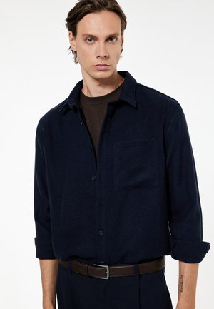 Jonge man met lichte huid en bruin haar, een donkerblauw overhemd met knopen over een zwart T-shirt, donkere broek en een bruine riem, staand tegen een witte achtergrond.