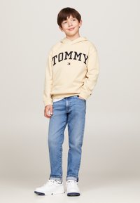 Felpa crema con "TOMMY" stampato in blu navy, abbinata a jeans azzurri. L'outfit include sneakers bianche con accenti blu navy.