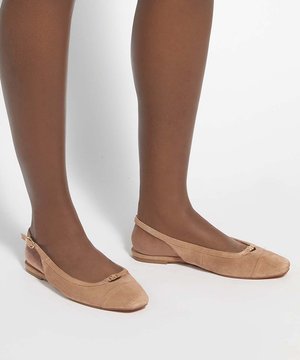 Brązowa skóra nóg nosząca beżowe zamszowe baleriny typu slingback z małą złotą klamrą na białym tle.