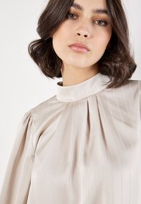 Blusa beige chiaro con righe verticali, collo alto e dettagli plissettati. Realizzata in tessuto morbido, presenta maniche a sbuffo.