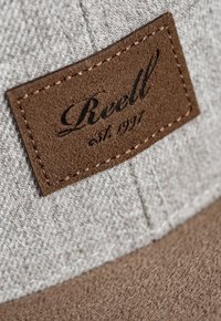 Gorra gris con una tela texturizada y un parche de ante marrón, que presenta el texto en relieve "Reell est. 1997" y detalles de costura en contraste.