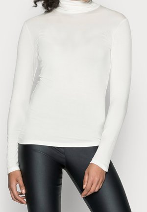 Henkilö, joka pukeutunut valkoiseen, istuvaan, pitkähihainen poolopaitaan ja mustiin nahkaa muistuttaviin leggingsseihin, seisoo rentoina käsin sivuilla.