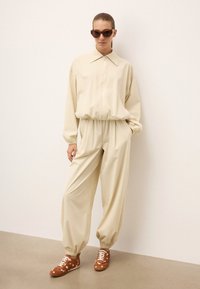 Beige zippinguga jope koos kraega, millele on sobitatud lõdvemat sorti beeži püksid. Pruunid tossud valgete täppidega komplimenteerivad kogu riietust.