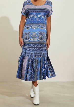 Robe bleue à motifs avec des manches courtes, arborant des designs floraux et des motifs paisley. Ourlet évasé avec un fond texturé. Portée avec des bottes de cowboy blanches.