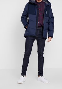 Laivastonsininen toppatakki vaaleansinisin yksityiskohdin, jossa on vetoketjullinen tasku ja huppu, yhdistetty tummansinisiin farkkuihin ja valkoisiin tennareihin.