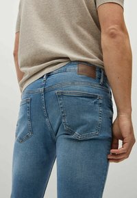 Jeans de mezclilla azul con corte slim, que presentan un parche de cuero marrón en la parte posterior, cinco bolsillos y un ligero desgaste cerca del bolsillo derecho.