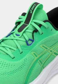 Tênis desportivo verde com parte superior em malha, superfície texturizada e sola de borracha. Apresenta atacadores, uma aba elástica para calçar e detalhes de marca em camadas.