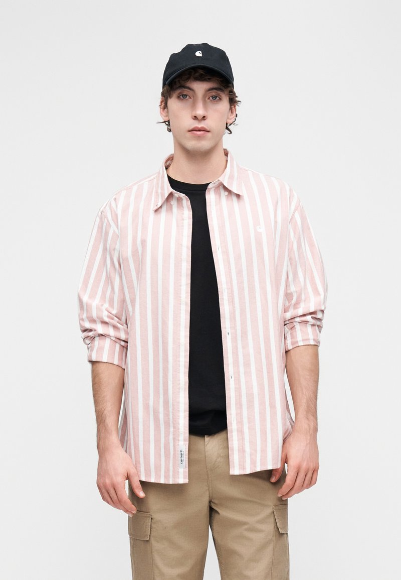 Chemise à manches longues rayée rose et blanche, boutonnée à l'avant, associée à un t-shirt noir et un pantalon cargo beige, casquette noire avec logo.
