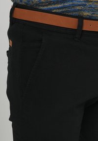 Svarta byxor i bomullsblandat tyg med slät textur, smal passform och ett brunt läderbälte som accentdetalj.