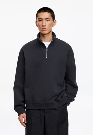 Jonge man met kort haar, draagt een donkerkleurige halve rits trui en een donkere broek, staand tegen een effen witte achtergrond.