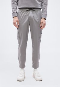 Άτομο που φοράει ανοιχτό γκρι παντελόνι cargo με κορδόνι στη μέση, λευκές κάλτσες και λευκά αθλητικά παπούτσια, στέκεται μπροστά σε απλό λευκό φόντο.