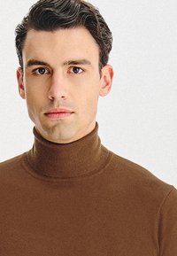 Brązowy sweter z golfem o prążkowanej fakturze, dopasowany krój i wysoki kołnierz. Gładki, bez wzorów i ozdób. Widoczna twarz modela.