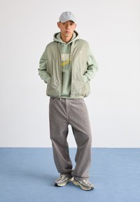 Hellgrüne, mit Reißverschluss versehene Weste über einem hellgrünen Hoodie mit gelbem Nike-Logo, kombiniert mit grauen Cordhosen und grauen Sneakern.