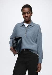 Sivý sveter zo zmesi vlny s golierovým výstrihom a štyrmi gombíkmi, v kombinácii s čiernymi džínsami, držiaci čiernu štruktúrovanú tašku.