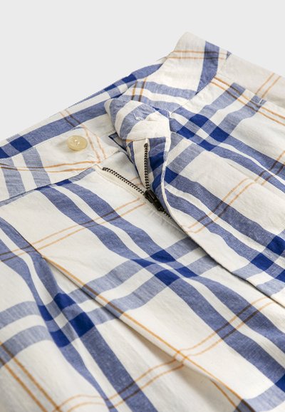 Tissu à carreaux bleu, blanc et beige avec une fermeture éclair partiellement ouverte et un bouton beige sur une ceinture de vêtement.