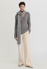 Grauer Strickpullover mit V-Ausschnitt und langen Ärmeln, kombiniert mit cremefarbenen, weit geschnittenen Hosen und einem langen grauen Schal mit pinken Akzenten.