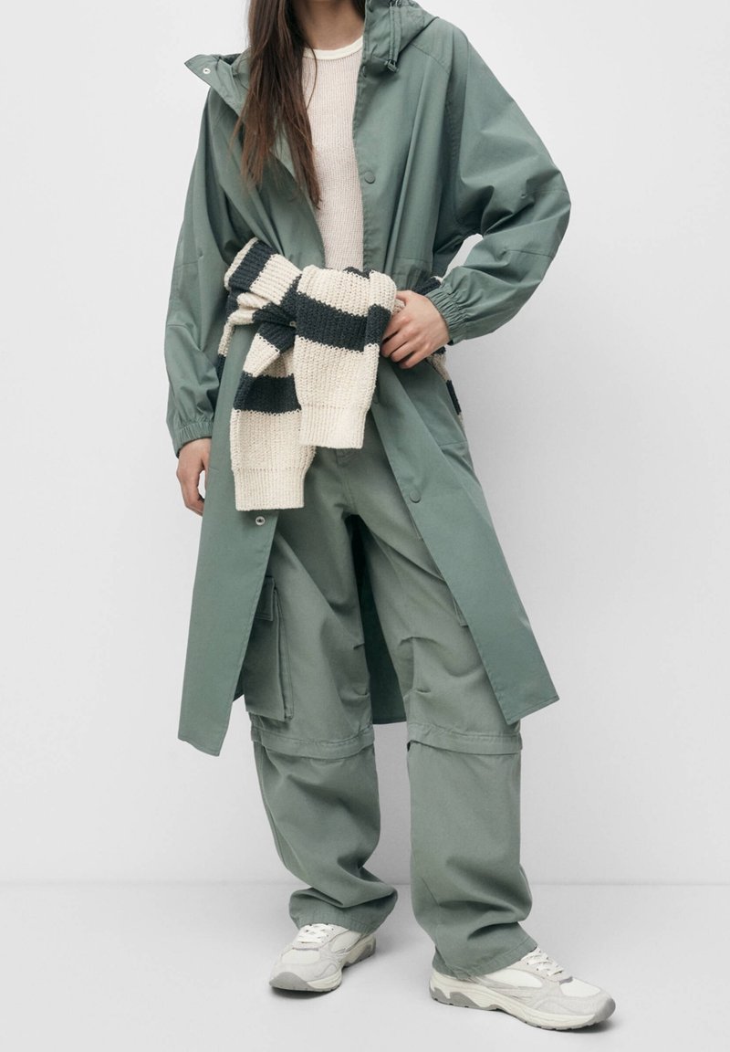 Personne portant un pantalon cargo ample vert sauge, un long imperméable assorti par-dessus un pull crème, avec un pull en tricot rayé noir et crème noué à la taille.