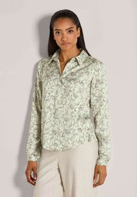 Vrouw in een lichtgroene blouse met paisley-patroon en lange mouwen, in combinatie met crèmekleurige broek, staand tegen een effen lichte achtergrond.