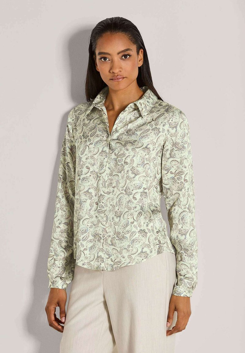 Vrouw in een lichtgroene blouse met paisley-patroon en lange mouwen, in combinatie met crèmekleurige broek, staand tegen een effen lichte achtergrond.
