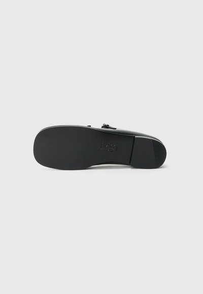 Svart skosula med en slät, platt design. Har en rundad kant och subtil prägling av logotypdetalj. Ren, minimalistisk estetik.