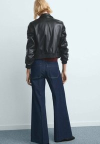 Personne aux cheveux blonds courts portant une veste en cuir noire, un jean large bleu foncé et des chaussures noires, debout contre un mur uni.