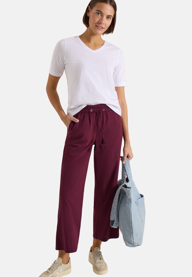Femme portant une chemise blanche à manches courtes et un pantalon large bordeaux, tenant un sac cabas bleu clair, debout devant un fond uni.