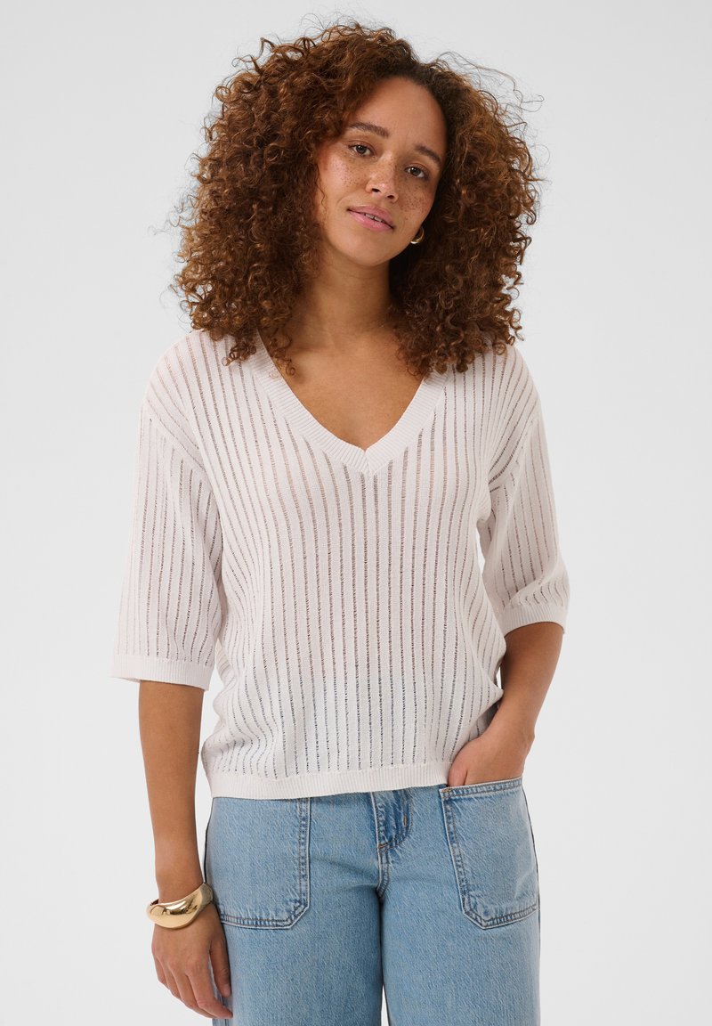 Femme aux cheveux bouclés porte un haut blanc, semi-transparent et rayé à col en V et un jean bleu clair, debout avec une main dans la poche.