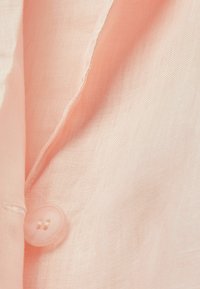 Světle růžová látka s měkkým, texturovaným povrchem, obsahující průhledný knoflík přišitý na okraji. Hladký, jemný detail tkaní je viditelný.